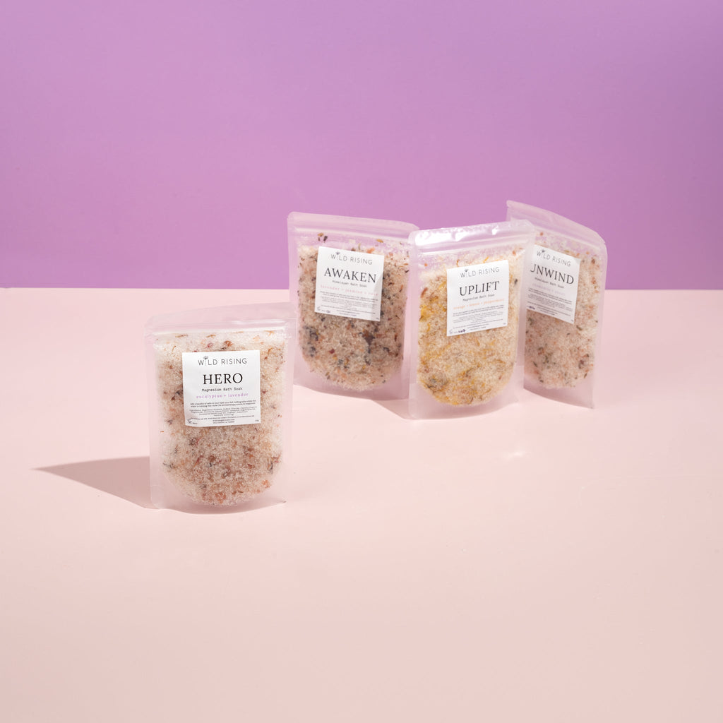 Four packages of the Bath Lovers Gift Set by WILD RISING SKINCARE lie on a pink surface with a purple background. Visible varieties are Hero, Awaken, Uplift, and Unwind—making this set perfect for relaxation and an ideal Christmas gift for someone special.