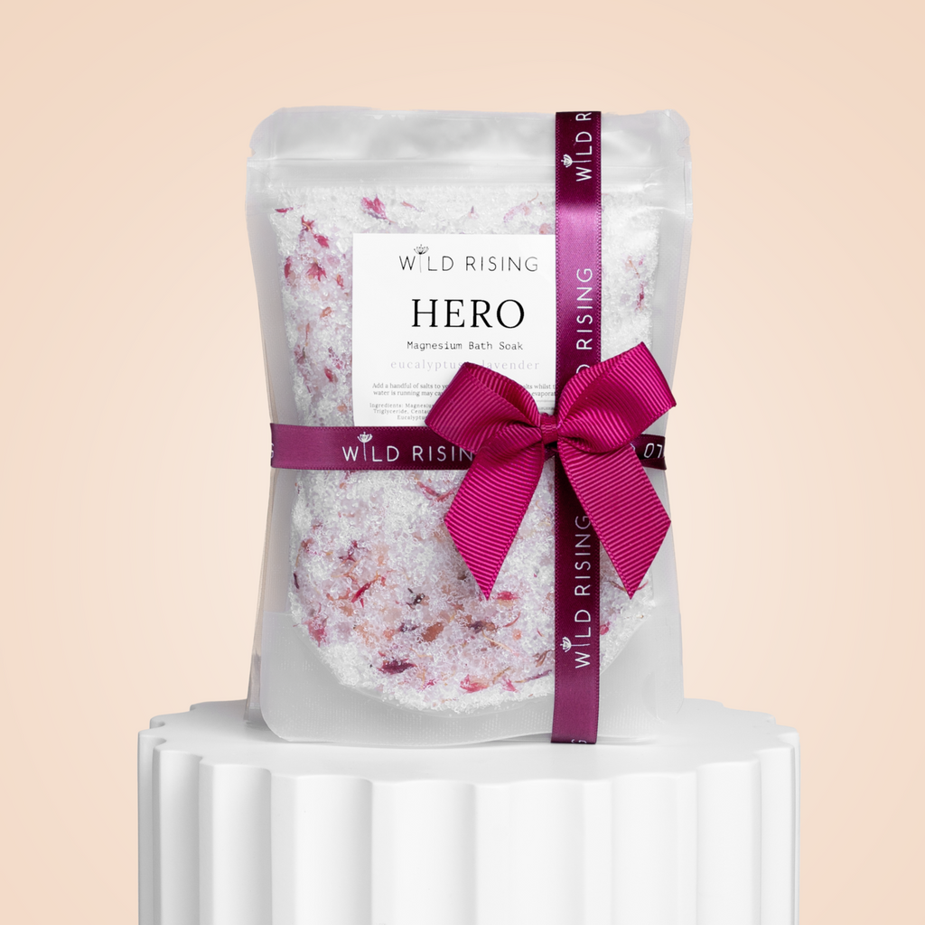 A clear WILD RISING SKINCARE Bath Lovers Gift Set rests elegantly on a white pedestal. Perfect for Christmas, it includes pink and white bath salts tied with a burgundy ribbon and branded tag, set against a soft peach background.