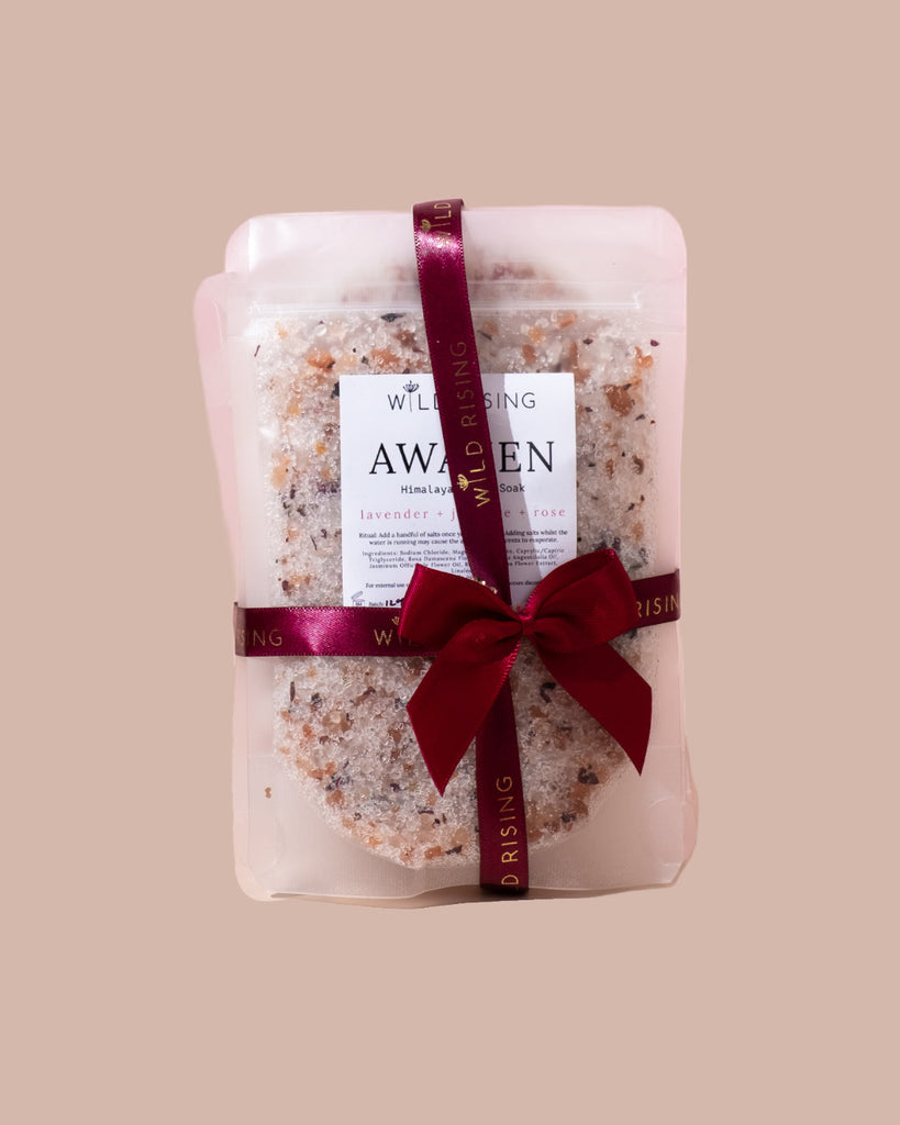A clear package of "AWAKEN" Himalayan bath salts from WILD RISING SKINCARE, tied with a red ribbon, is a perfect Christmas gift. Infused with lavender and rose, this bath set makes a charming stocking filler against its plain beige packaging.