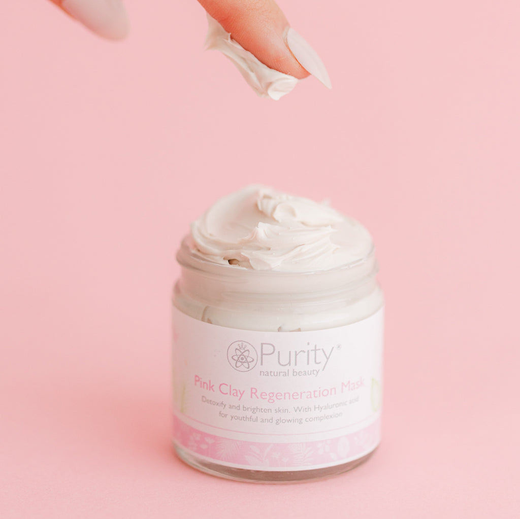 A manicured hand scoops creamy pink clay from a PurityNaturalBeautyUK jar labeled "Pink Clay Face Mask," set against a soft pink backdrop. Enriched with natural ingredients, this mask promises to detoxify the skin for a youthful, glowing radiance.